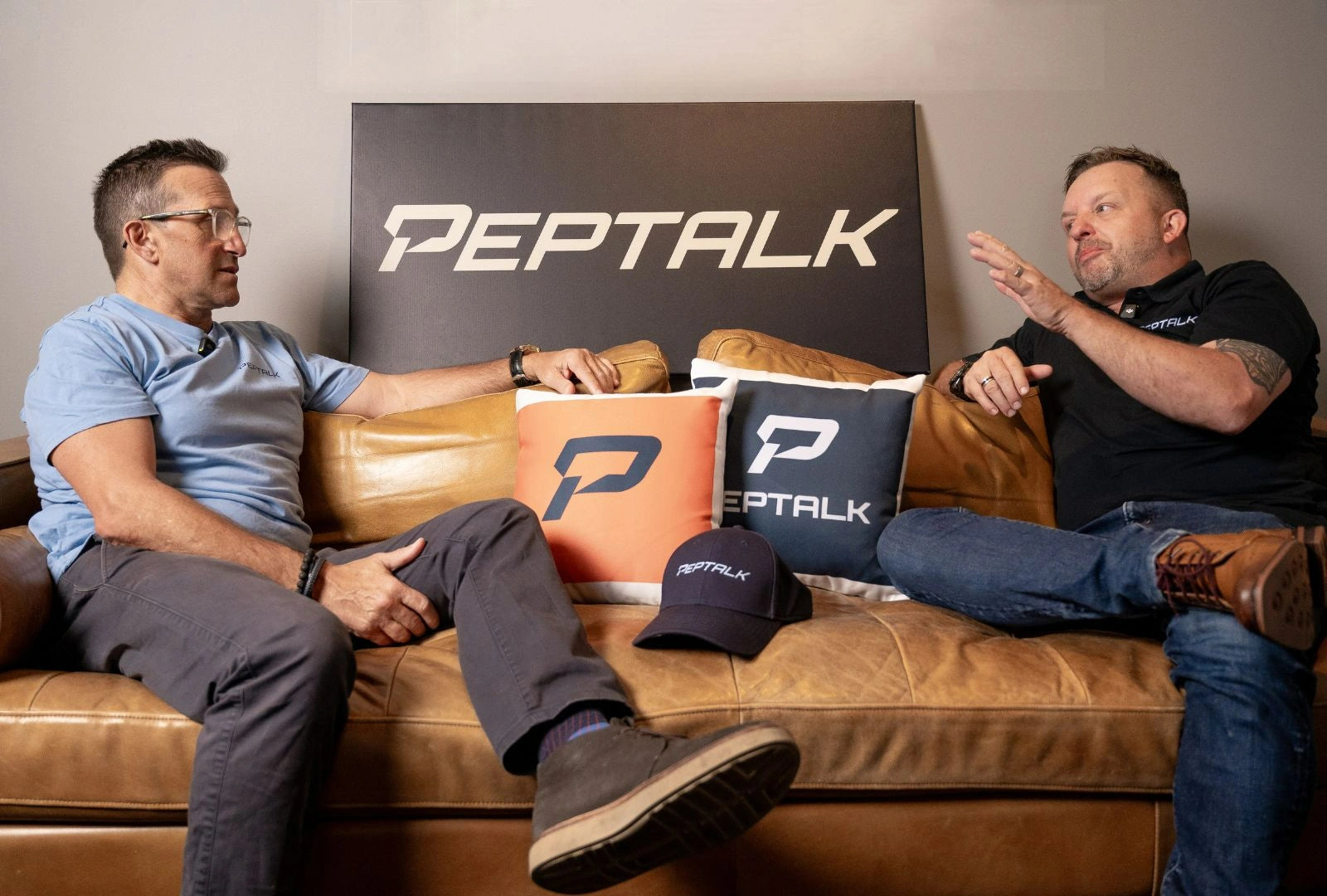 Photo of Matt and Pepe talking while sitting on a leather couch, with some PepTalk branded items around: two pillows, a cap and a signage