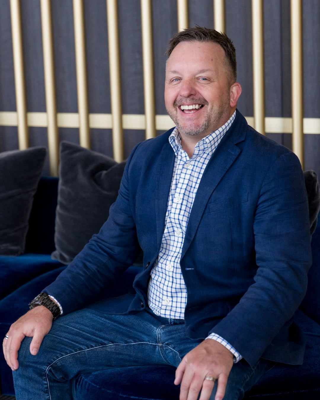 Photo of Matt Clayton smiling and wearing a checkered polo and blue coat while sitting on a couch